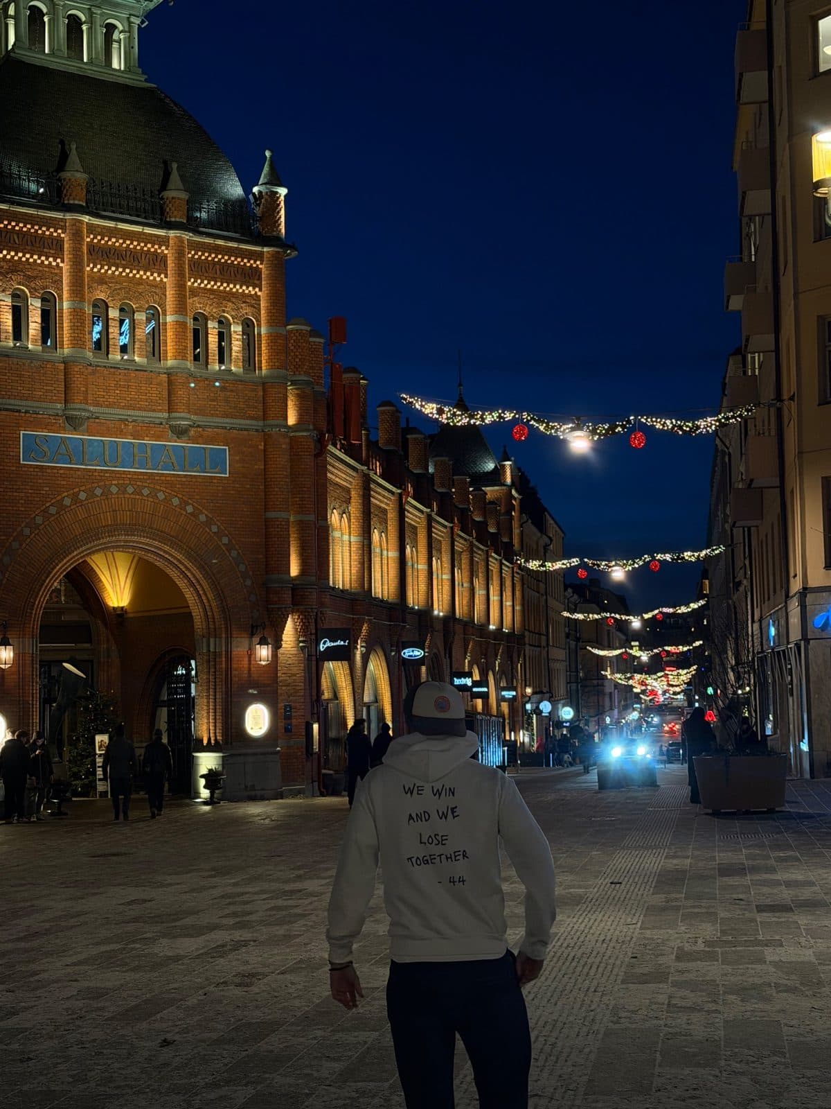 A person walking down an evening street in a white Scudaro hoodie with the back print “We win and we lose together – 44”
